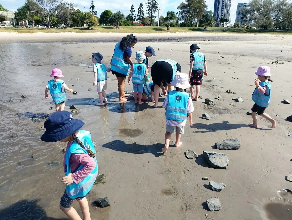 Children in high-visibility vests and sun hats exploring the wet sand and shallow water at the shoreline during a Beach Kindy session, with an educator alongside them