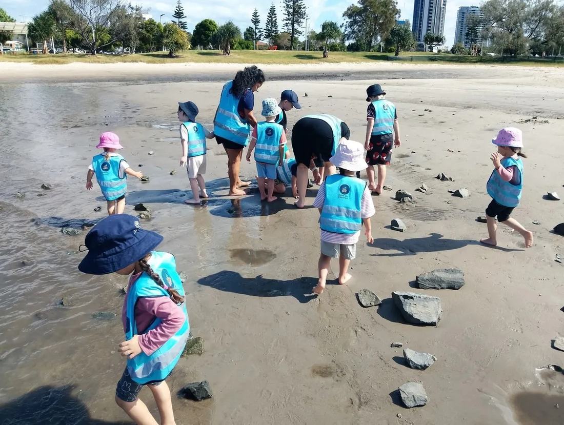 Children in high-visibility vests and sun hats exploring the wet sand and shallow water at the shoreline during a Beach Kindy session, with an educator alongside them